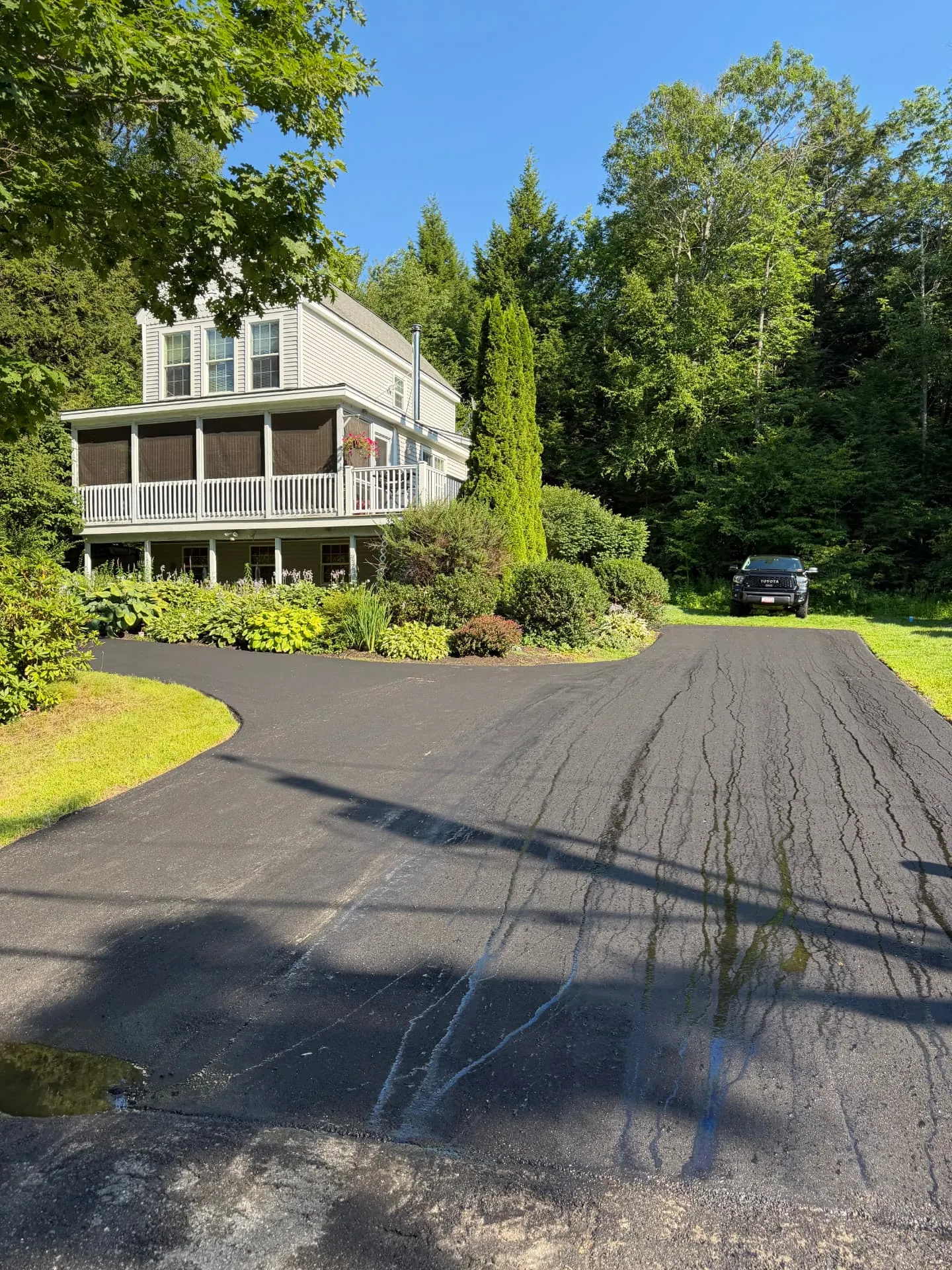 Freshly paved curved asphalt driveway leading up to a Maine home with gardens and a pickup truck