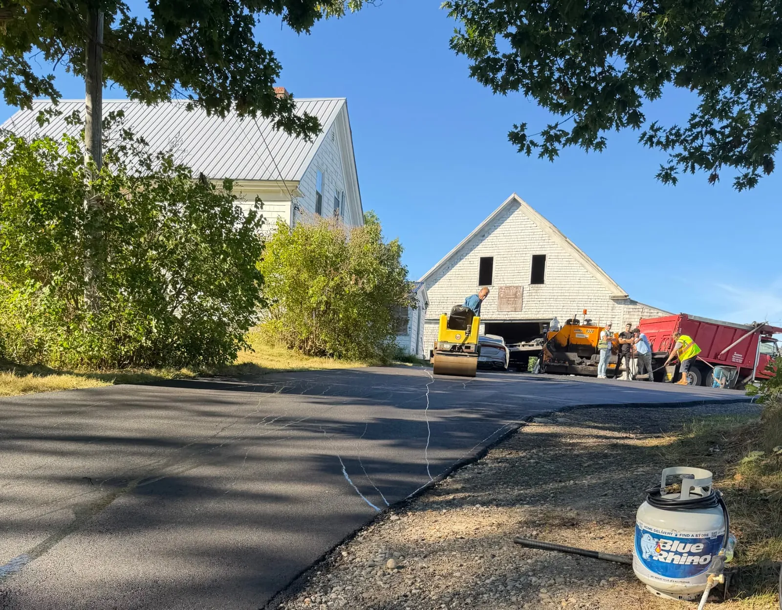Paving crew with paver, roller and dump truck at a white farmhouse barn